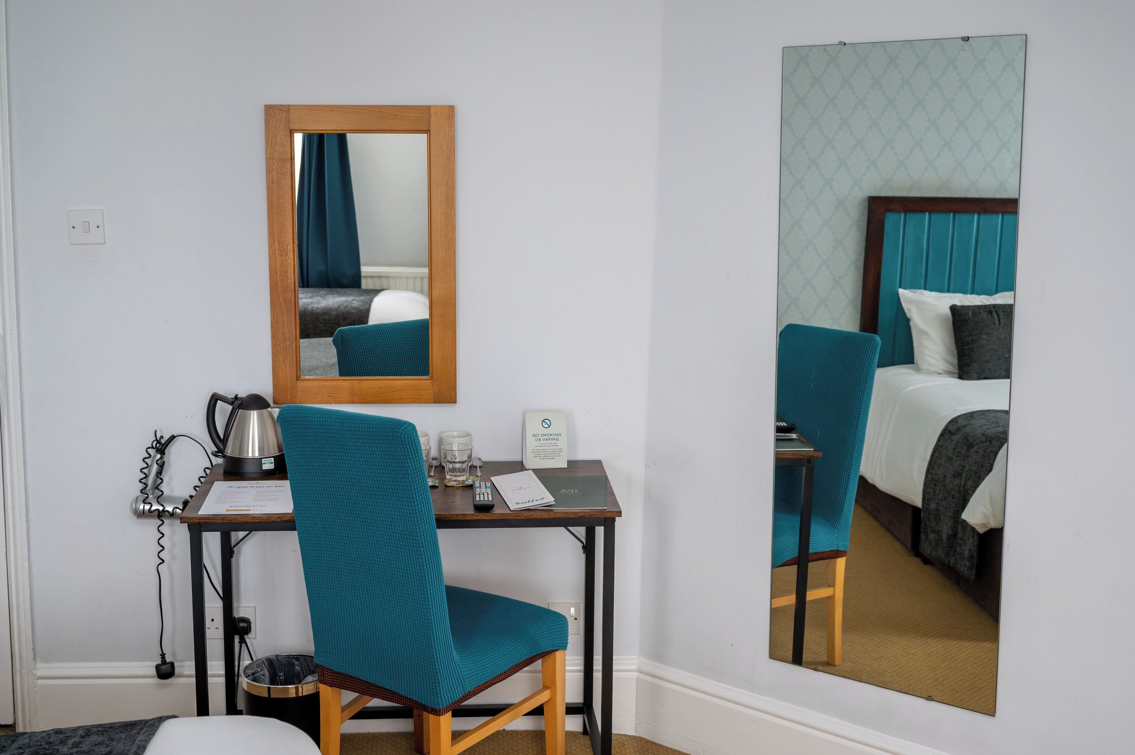 A hotel room with a wooden desk, a teal chair, a bed with teal headboard, a mirror, and a "No Smoking" sign.