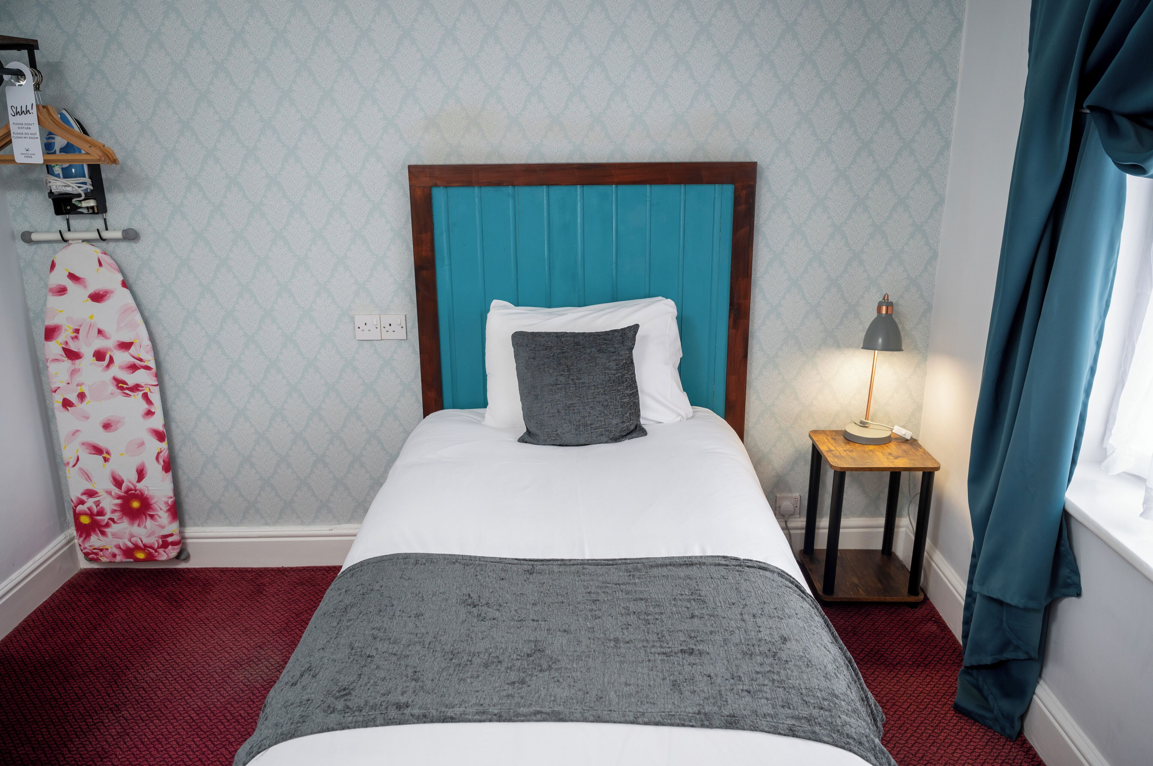 Una cama bien hecha con un cabecero verde azulado, una almohada gris y una almohada blanca. Una mesita de noche de madera con una lámpara está al lado de la cama. Un tabla de surf cuelga en la pared a la izquierda.