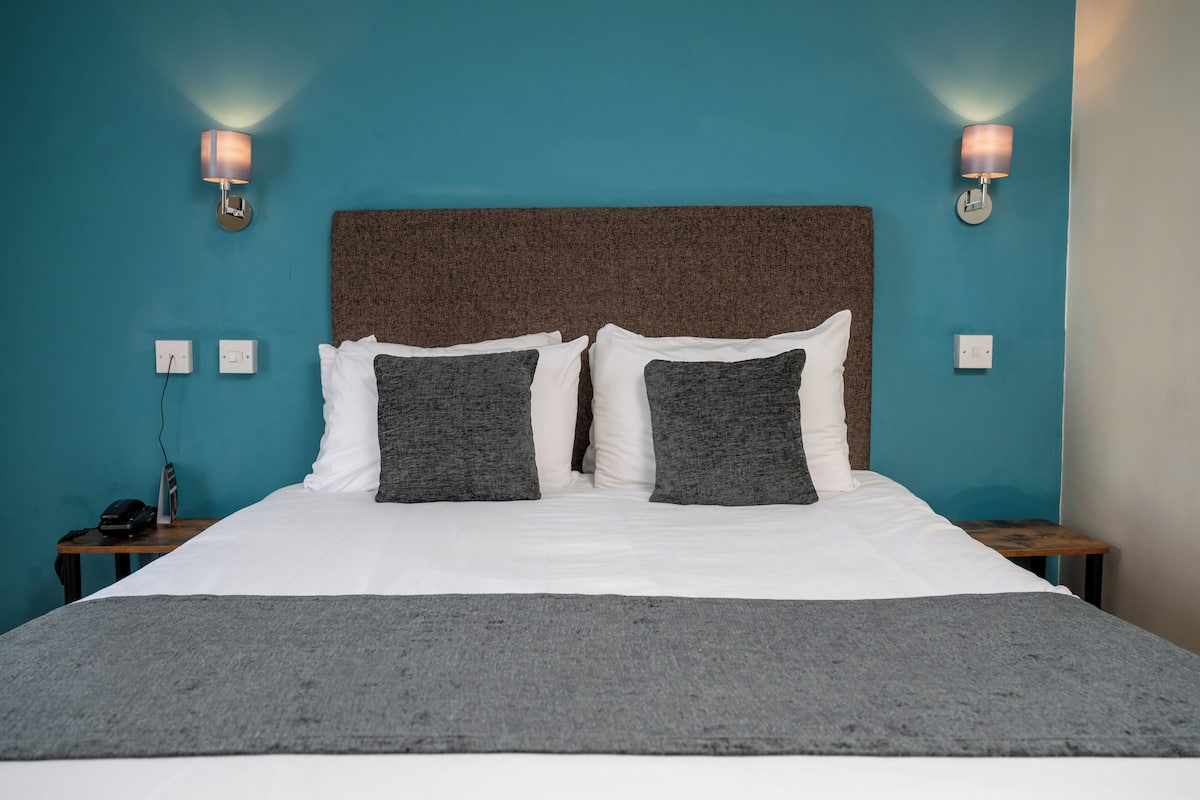 A neatly made bed with a grey blanket, two pillows, and a brown headboard. There are two bedside tables with lamps and a phone on one.