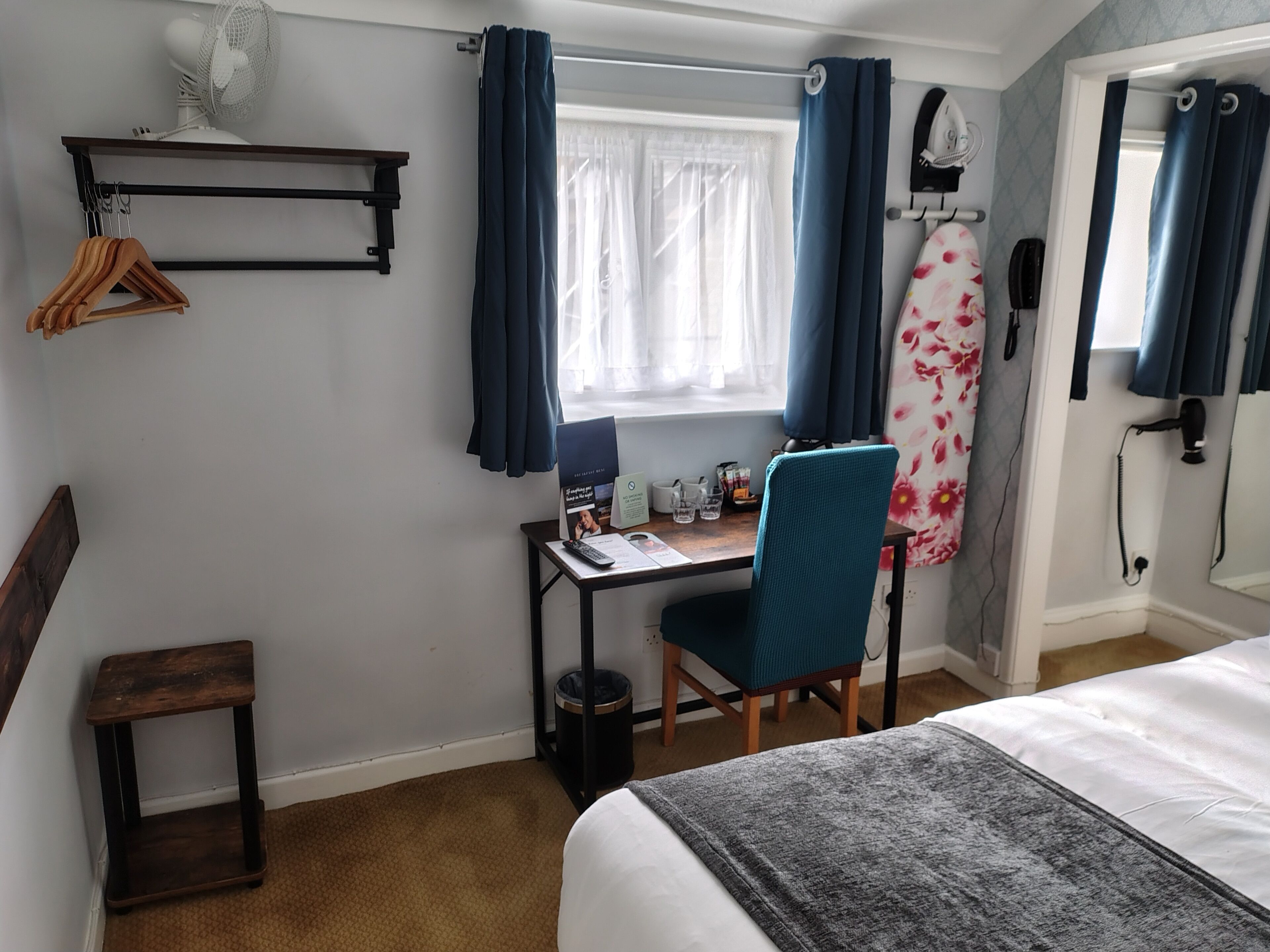 Habitación de hotel con cama, escritorio y silla, una mesita, perchero, ventana con cortinas, espejo y secador de pelo.