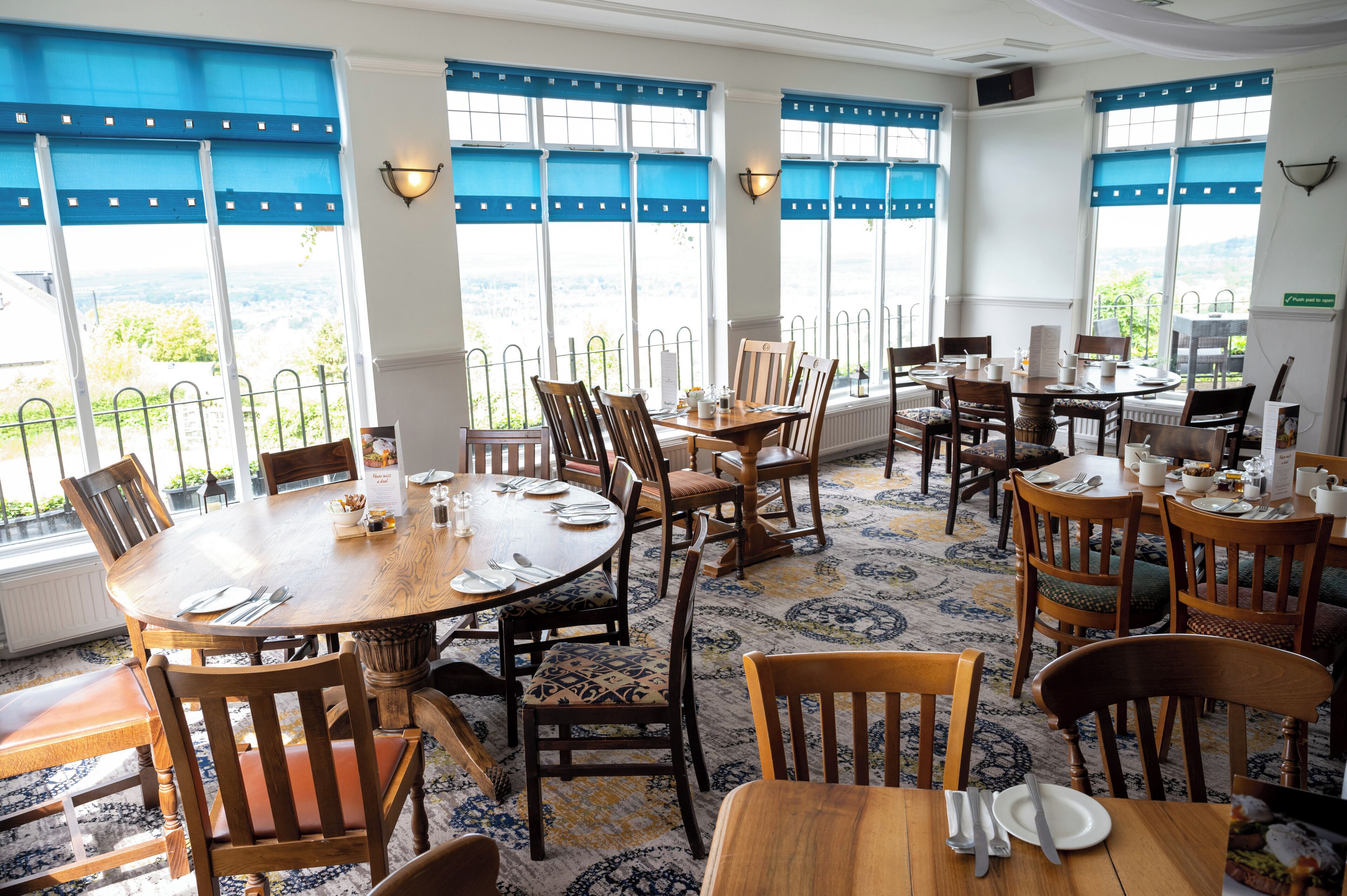 Un espace repas avec des tables et des chaises rondes, de grandes fenêtres équipées de stores bleus et une vue sur l’extérieur.