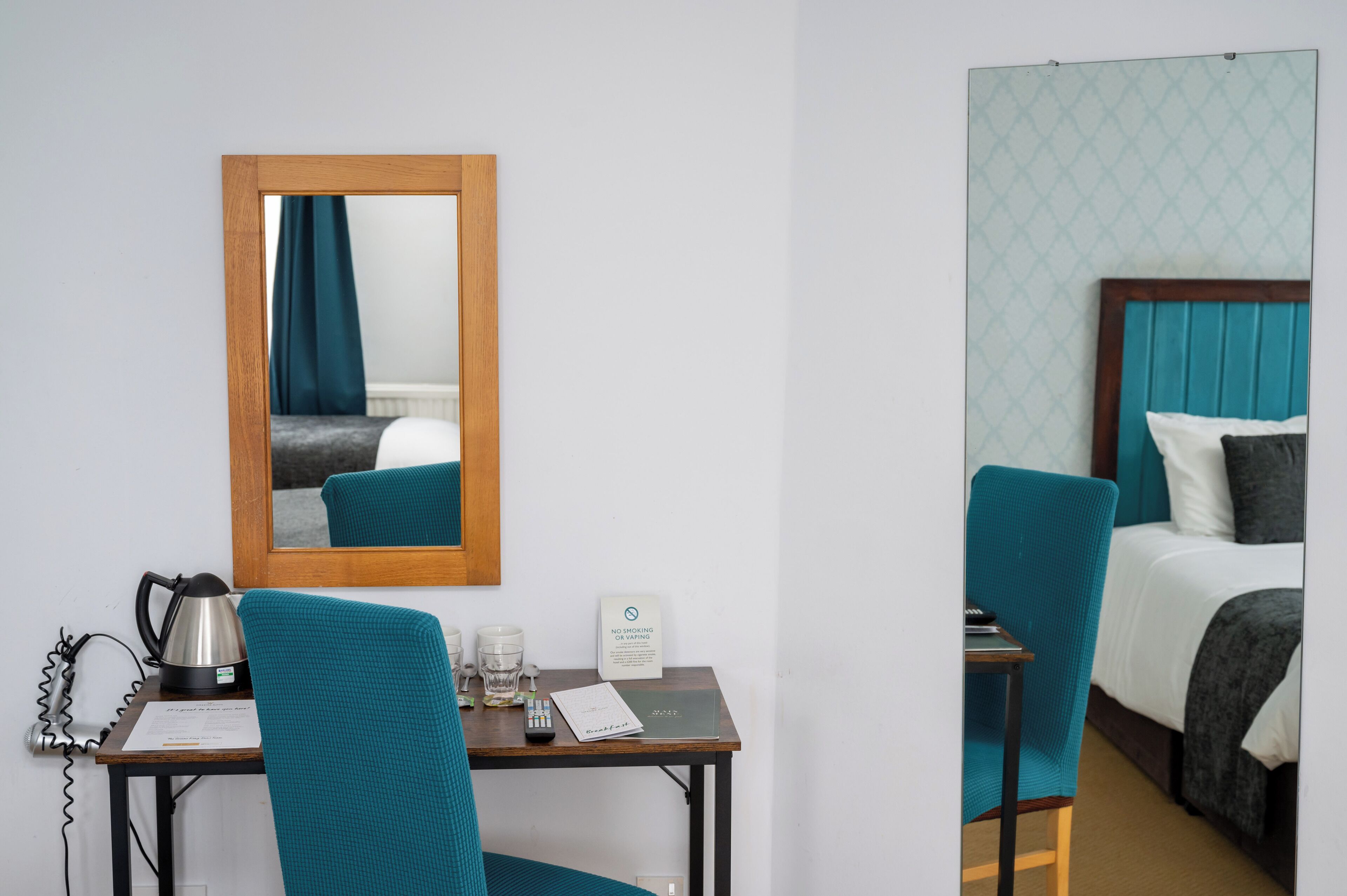 Habitación de hotel con espejo de marco de madera, una silla color turquesa, una mesita con hervidor, vasos, un cartel y una cama con cabecero y almohadas color turquesa.