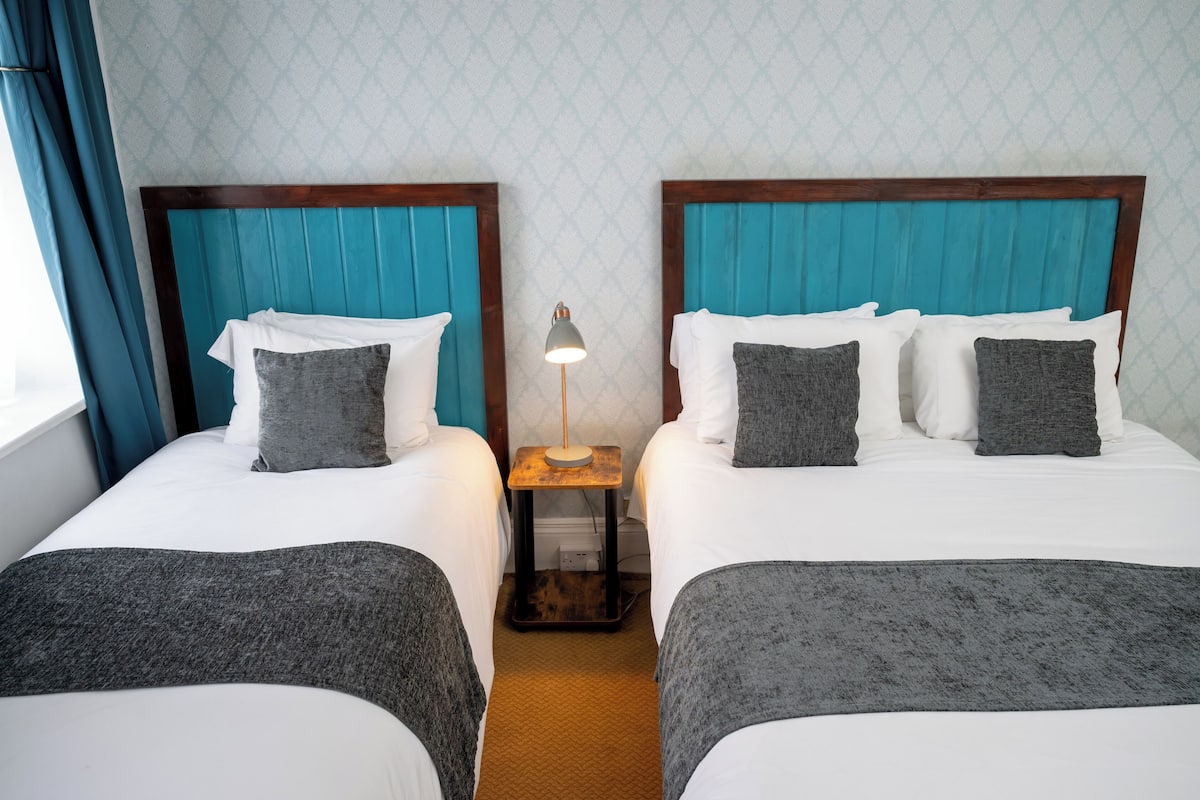 A hotel room with two beds, each with a wooden headboard and a teal accent. There is a small table with a lamp between the beds.