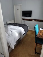 A single bed with white and grey bedding, a television mounted on the wall, a wooden headboard, and a small table with a chair.