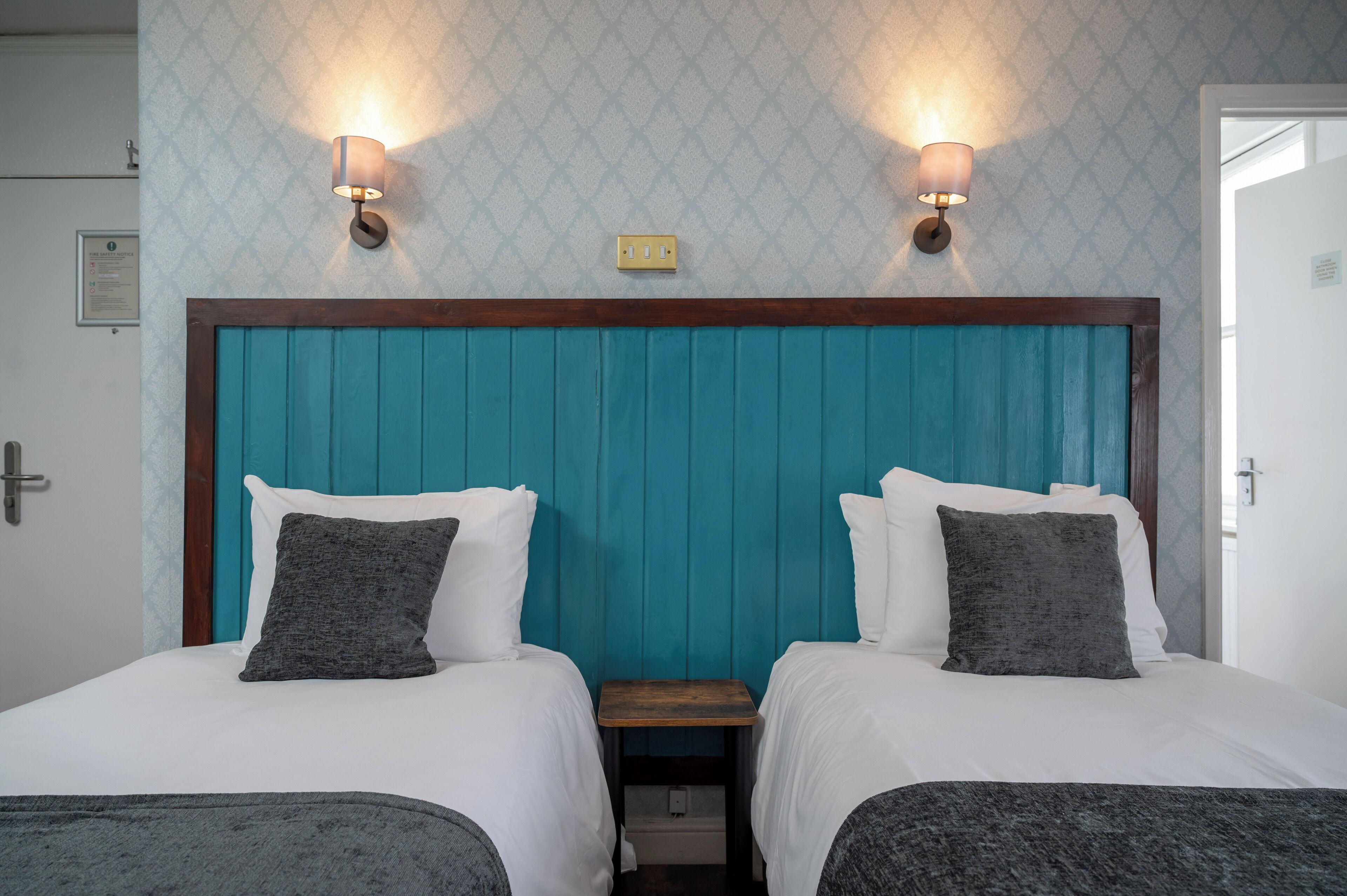 Habitación de hotel con dos camas, cabecero color turquesa, ropa de cama blanca e iluminación montada en la pared.