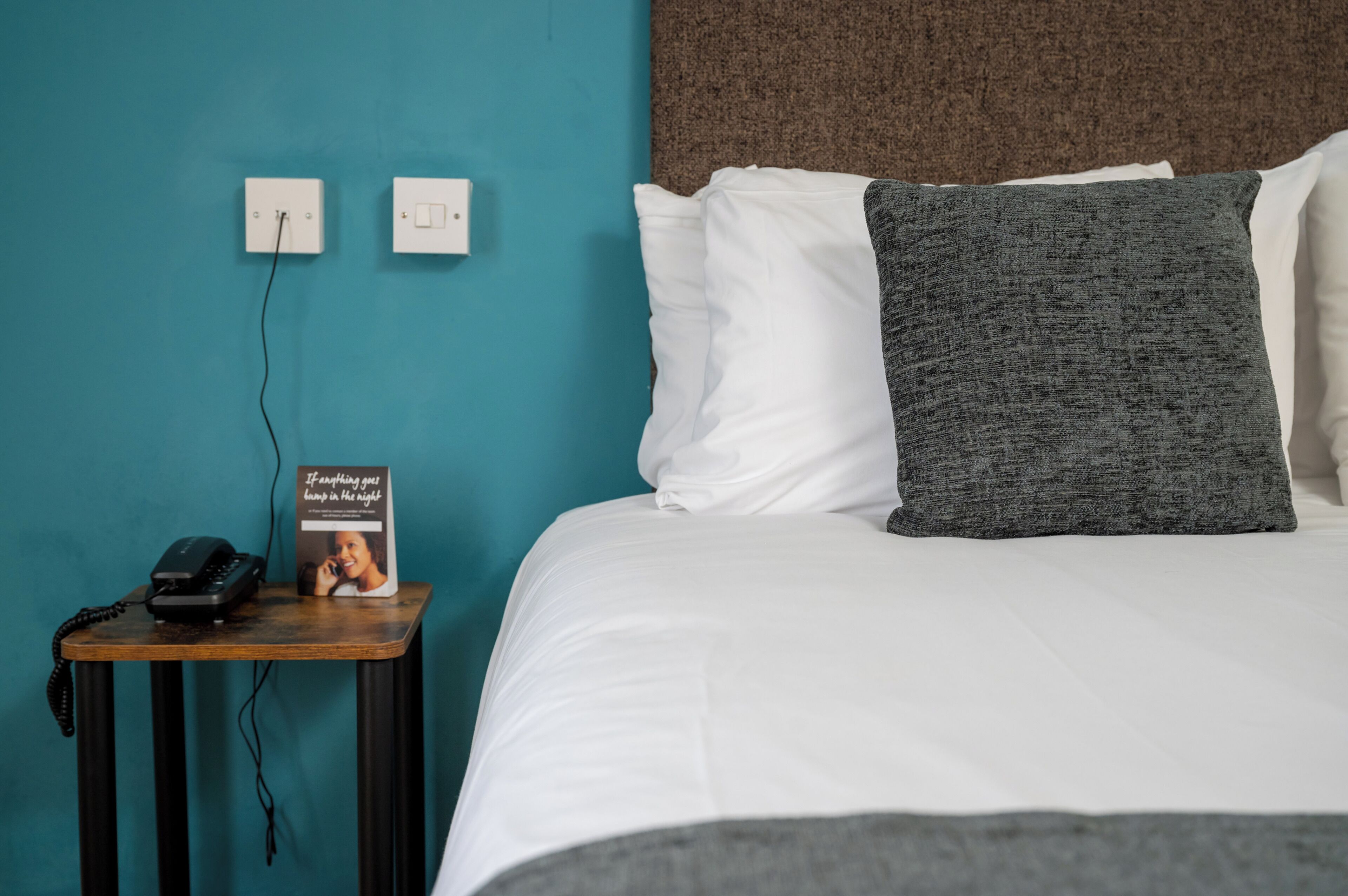 A neatly made bed with white and gray pillows, a wooden nightstand with a telephone and a brochure, and two wall switches on a teal wall.