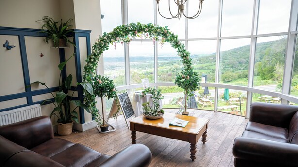 A room with a view of a green landscape, a wooden table, and leather sofas. - Rising Sun Hotel by Greene King Inns (Cheltenham)