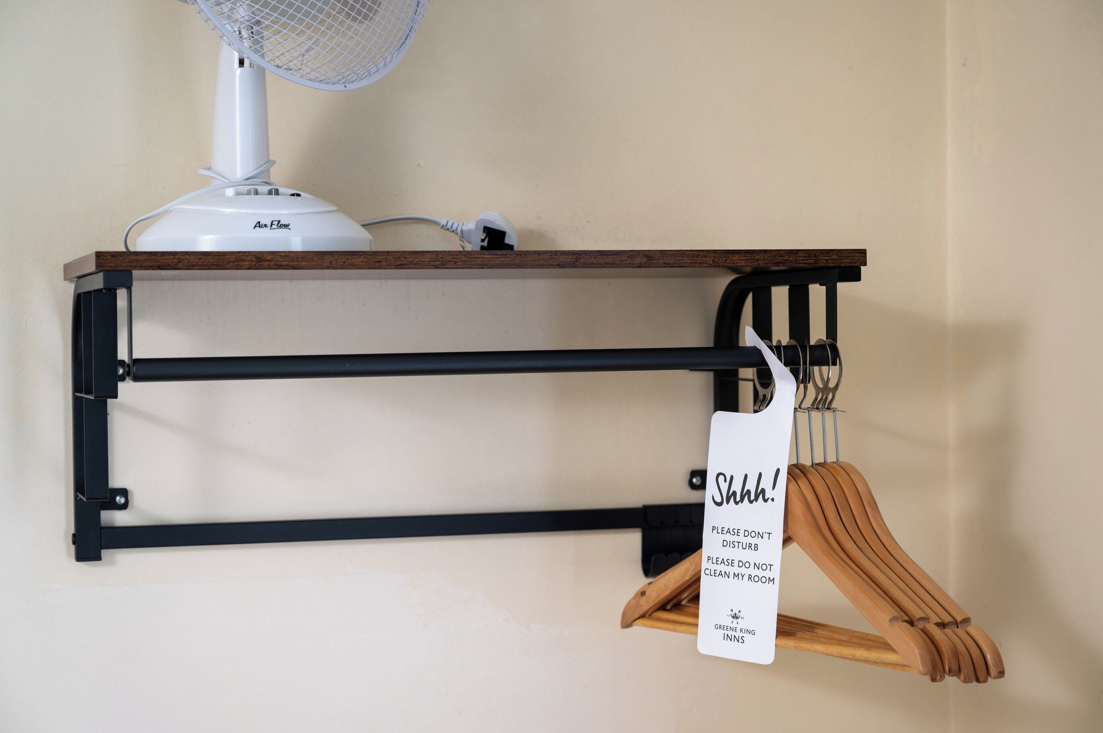 A wall-mounted shelf with a white fan, a wooden hanger rack, and a sign with a message.