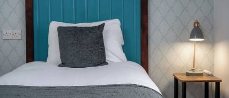 Una cama bien hecha con un cabecero verde azulado, una almohada gris y una almohada blanca. Junto a la cama, una mesita de noche de madera con una lámpara.