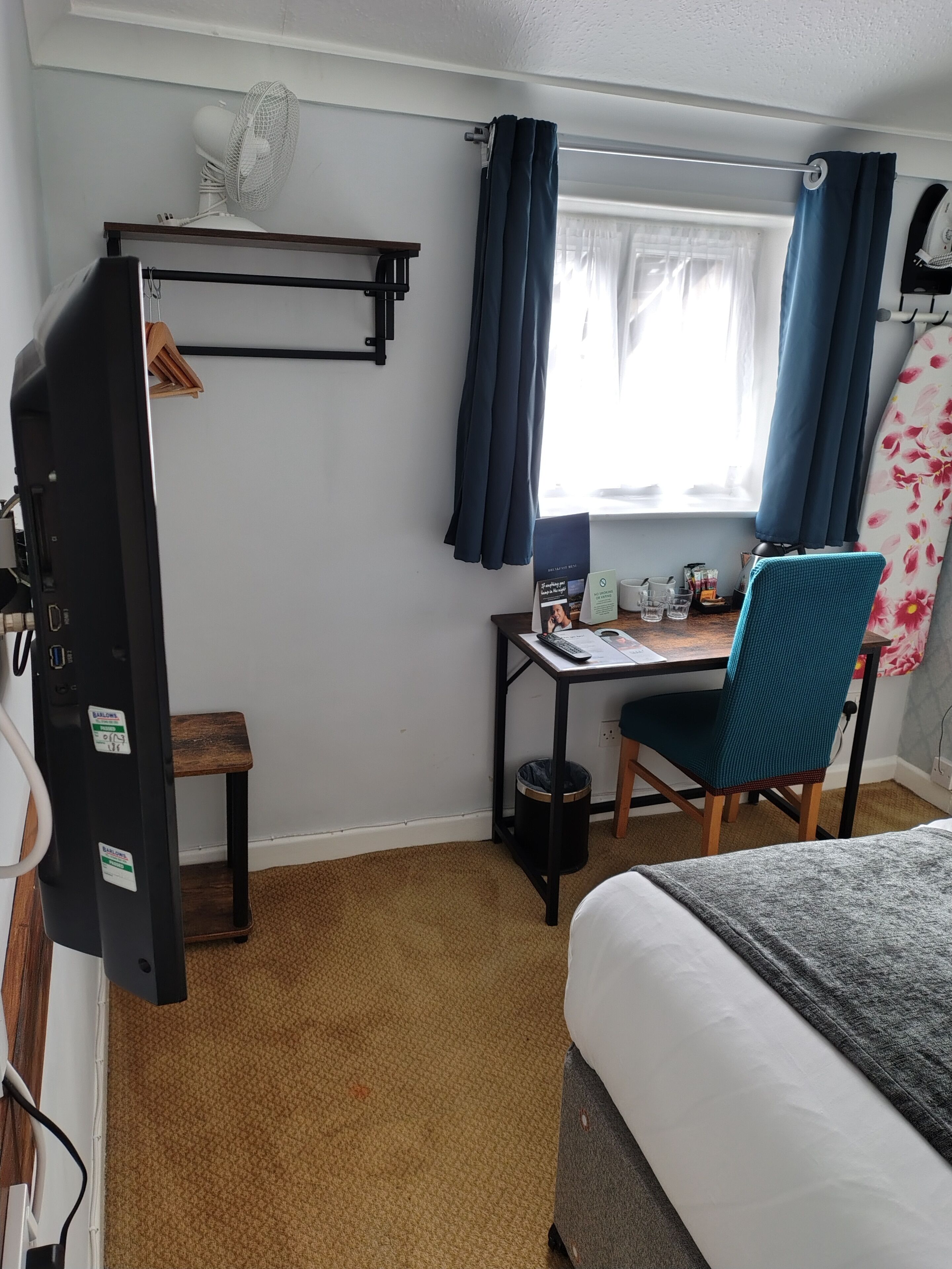 Habitación de hotel pequeña con cama, escritorio con silla, una mesita, ventilador y ventana con cortinas.