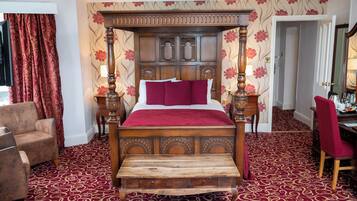 A four-poster bed with a wooden headboard, a red bedspread, and a red pillow. There is a wooden bench at the foot of the bed. The room has a red carpet and floral wallpaper.