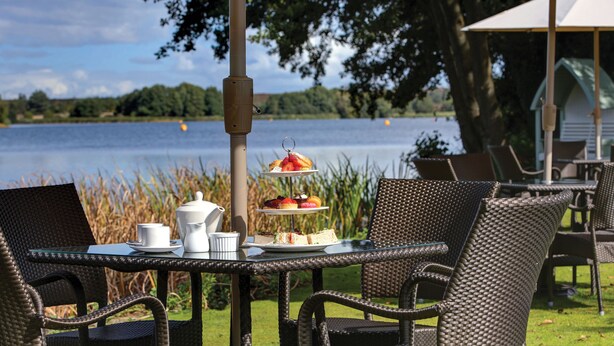 An outdoor dining area with wicker chairs and a table set for a meal, overlooking a lake. - Frensham Pond Country House Hotel & Spa (Farnham)