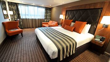 A hotel room with a large bed, two armchairs, a nightstand with a lamp, and a view of the outdoors through curtains.