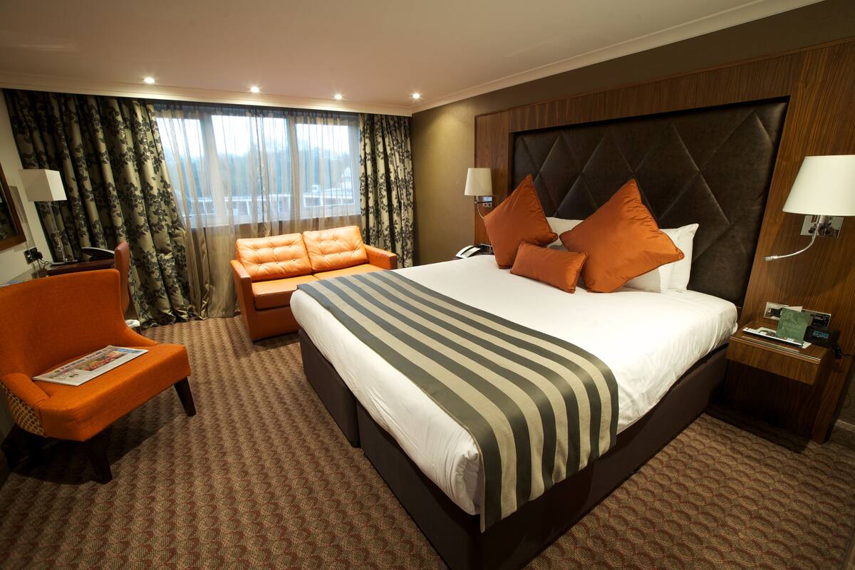 A hotel room with a large bed, a sofa, a chair, and a nightstand with a lamp.