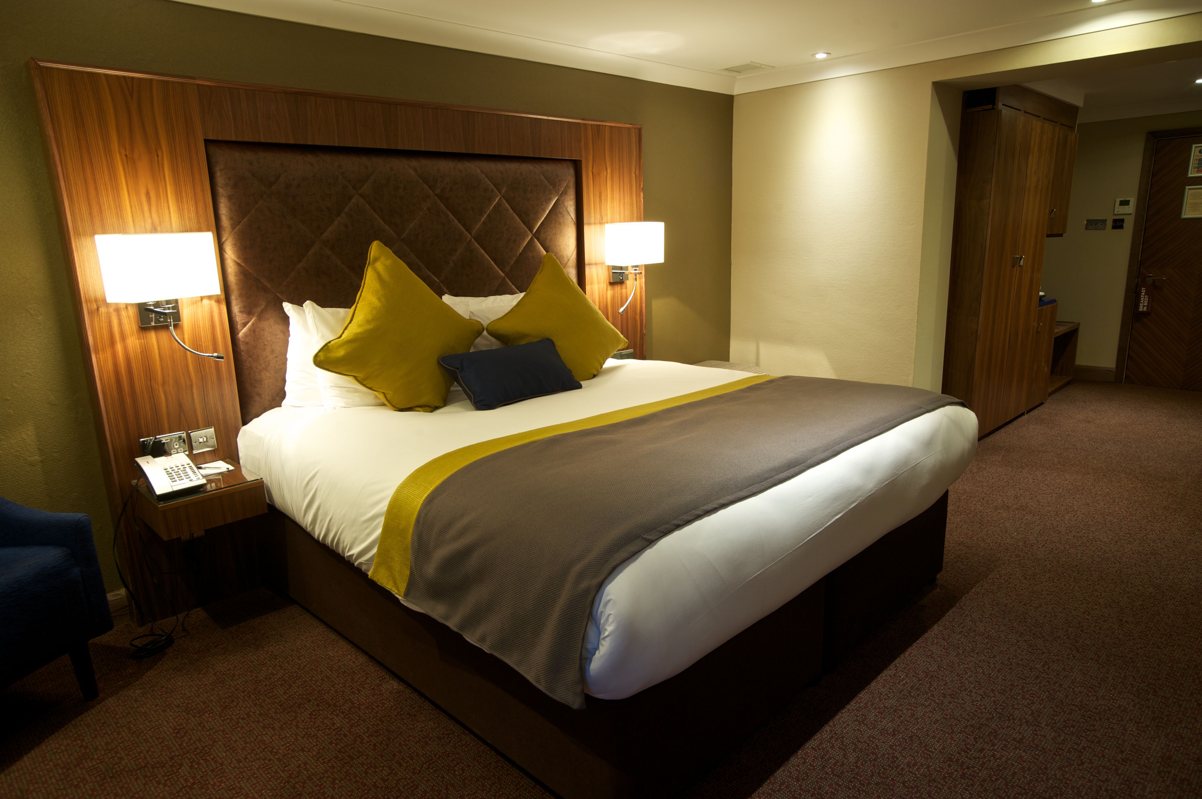 A hotel room with a large bed, two bedside lamps, a wooden headboard, and a desk with a telephone.