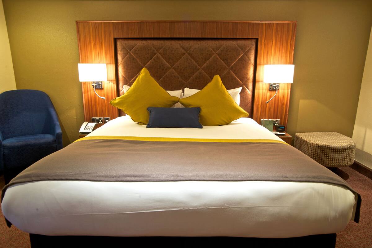 A large bed with a quilted headboard, two bedside lamps, a blue chair, and a small ottoman.
