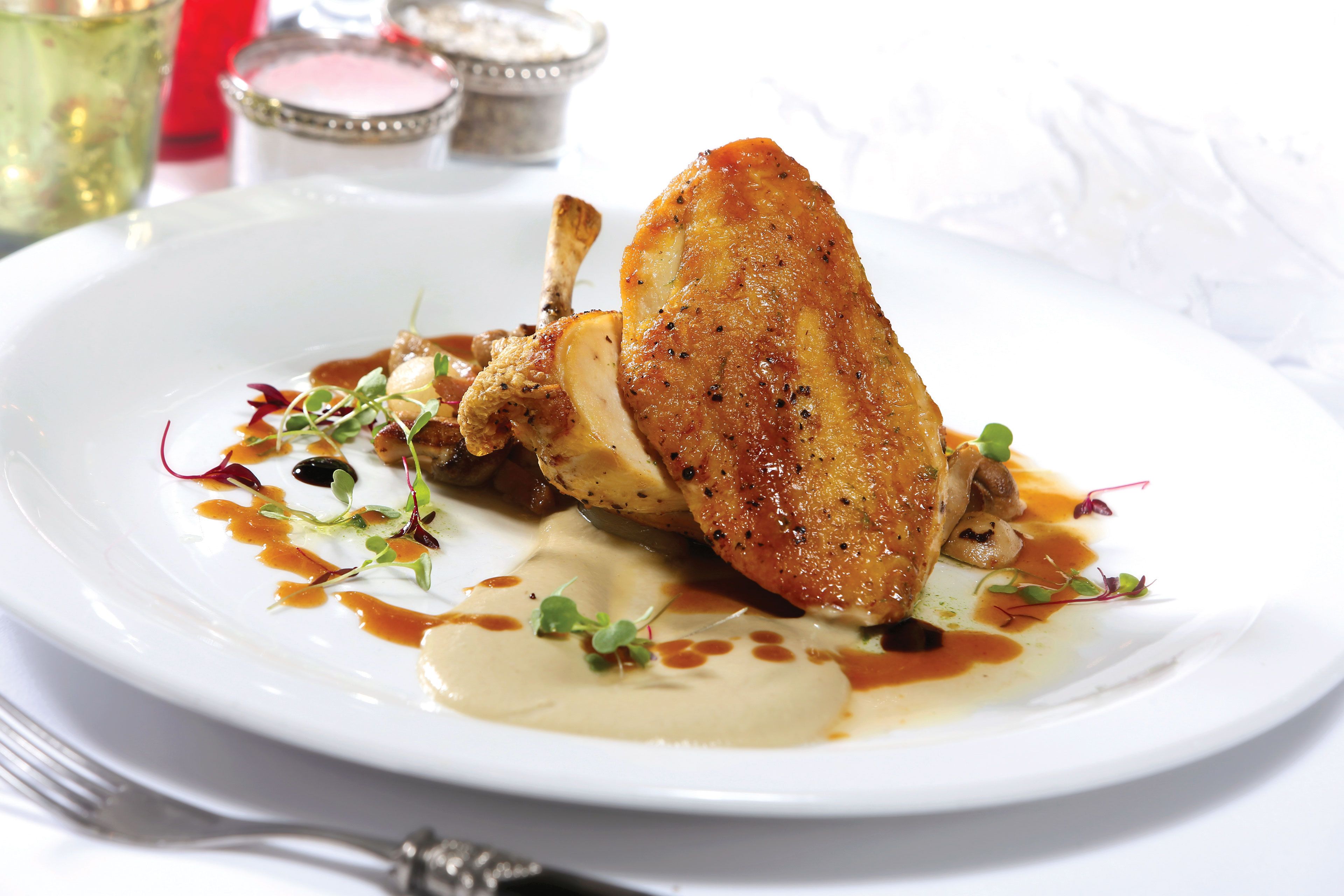 Un plato blanco con un muslo de pollo asado, salsa y guarniciones.