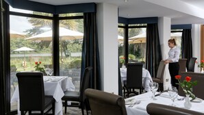A restaurant with tables set for dining, large windows with curtains, and a view of a lake. - Frensham Pond Country House Hotel & Spa (Farnham)