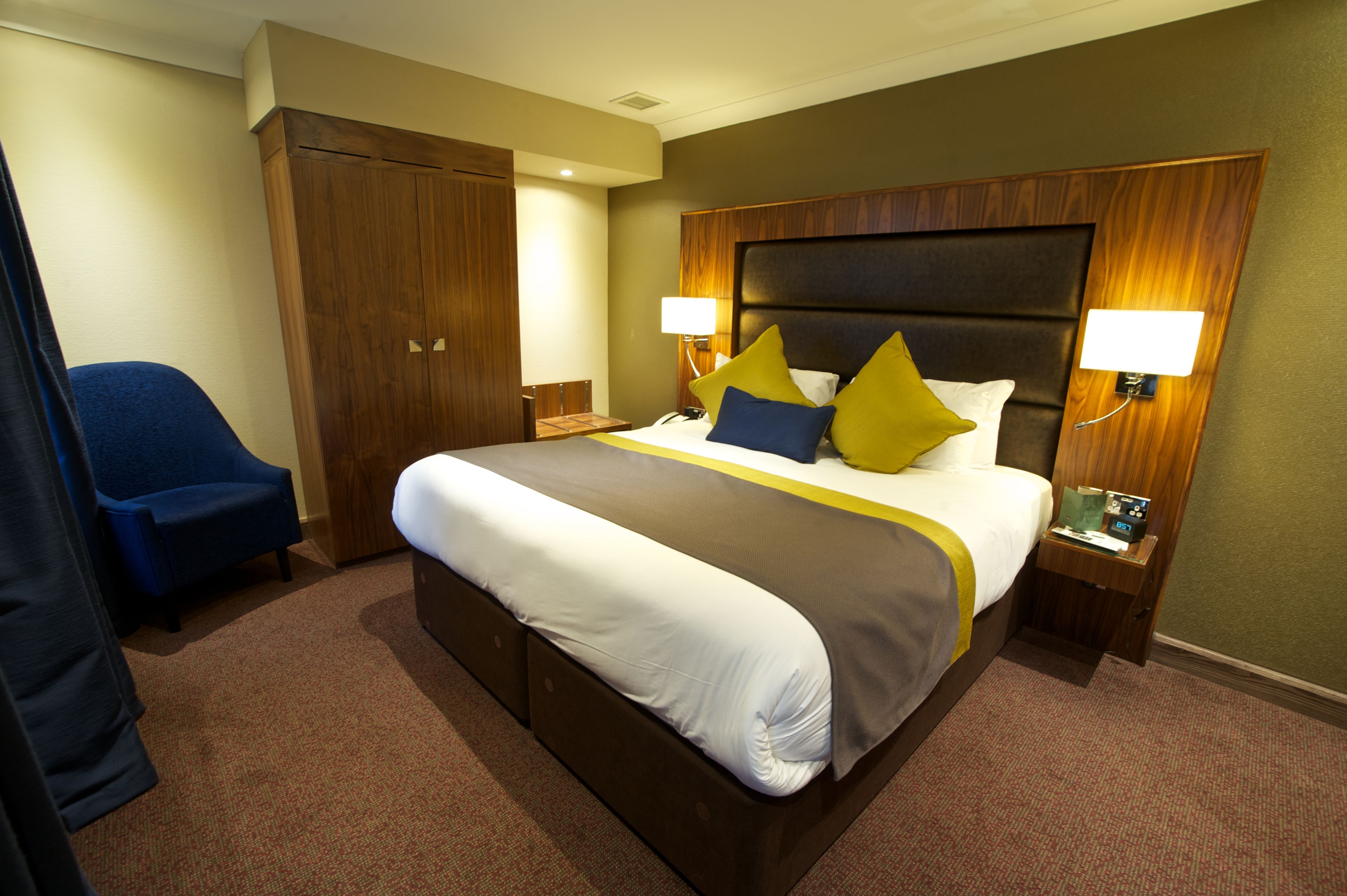 Habitación de hotel con una cama grande, dos lámparas de noche, un cabecero de madera, un sillón azul y un armario.
