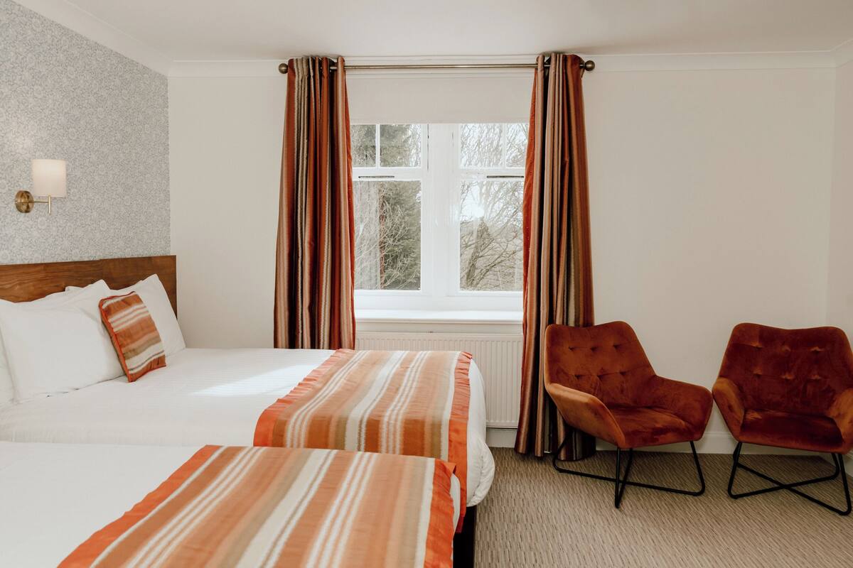 Une chambre d’hôtel avec deux lits, une fenêtre avec des rideaux, une lampe et deux fauteuils.