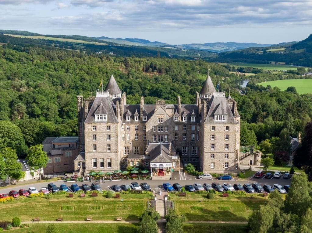 A large castle-like hotel with multiple towers, a parking lot, and a view of the surrounding green landscape.