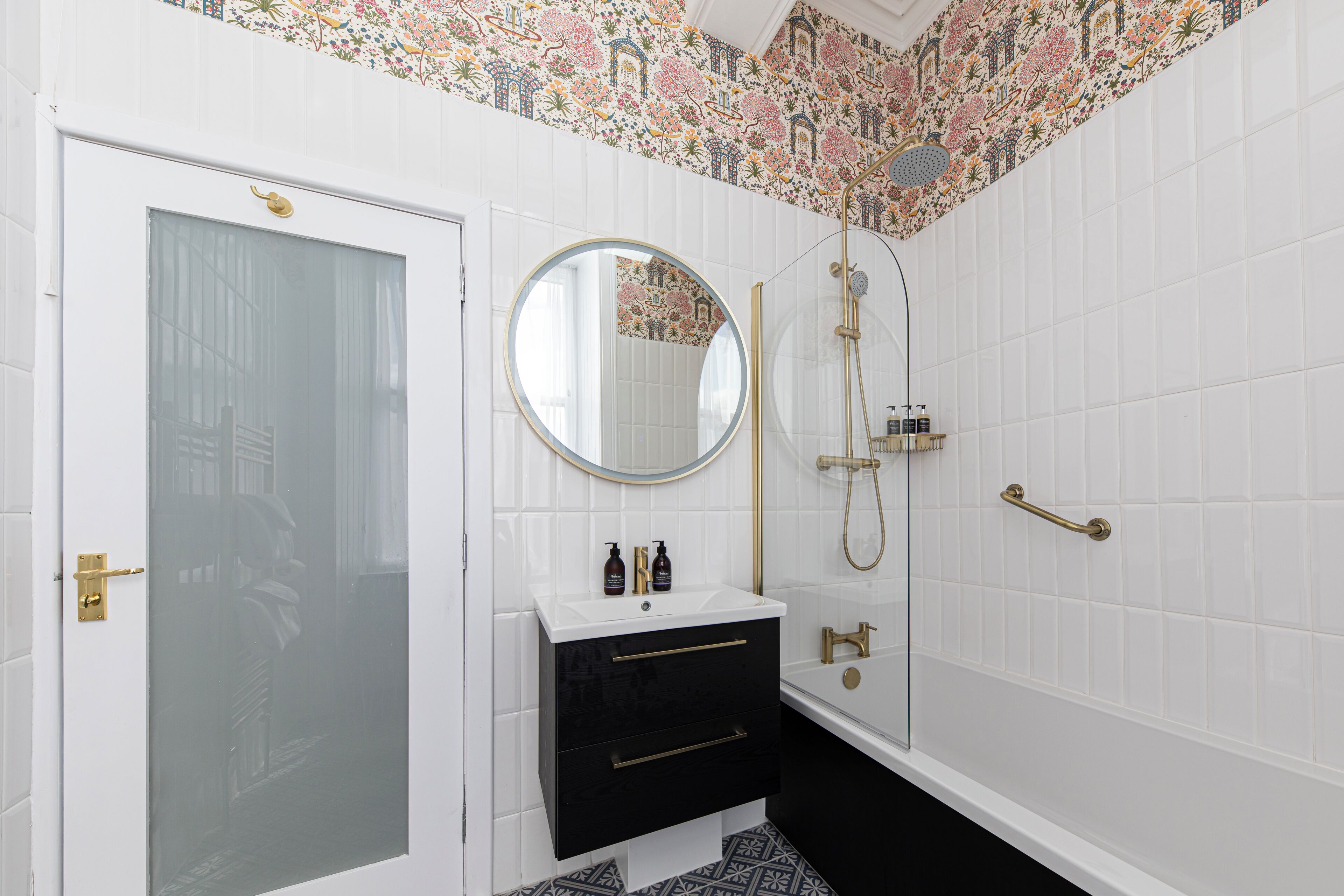 A bathroom with a white tiled wall, a black vanity with gold handles, a round mirror, and a shower area.