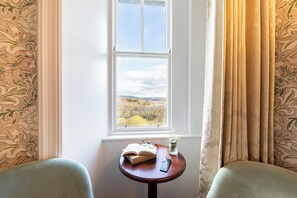 A room with a window offering a view of a landscape, a small table with a book, glasses, and a vase with flowers. - Atholl Palace Hotel (Pitlochry)