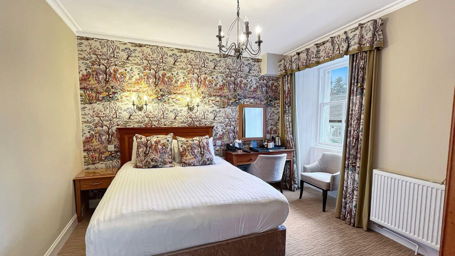 A hotel room with a large bed, a desk, a chair, a chandelier, and a window with curtains.
