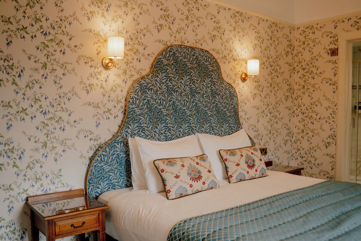 Une chambre à coucher avec un papier peint à motifs floraux, une tête de lit, deux lampes de chevet, une table de chevet en bois et un lit agrémenté d’oreillers à motifs floraux.