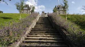 A stone staircase leading to a large stone building with multiple chimneys, surrounded by greenery and lavender bushes.