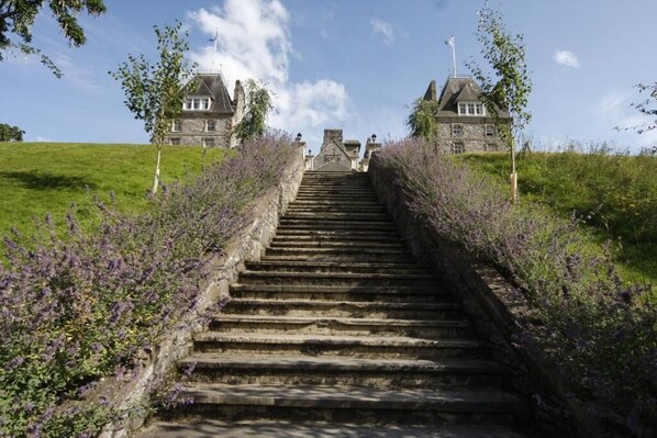 Una escalera de piedra conduce a un edificio grande de piedra con múltiples chimeneas, rodeado de follaje y arbustos de lavanda.