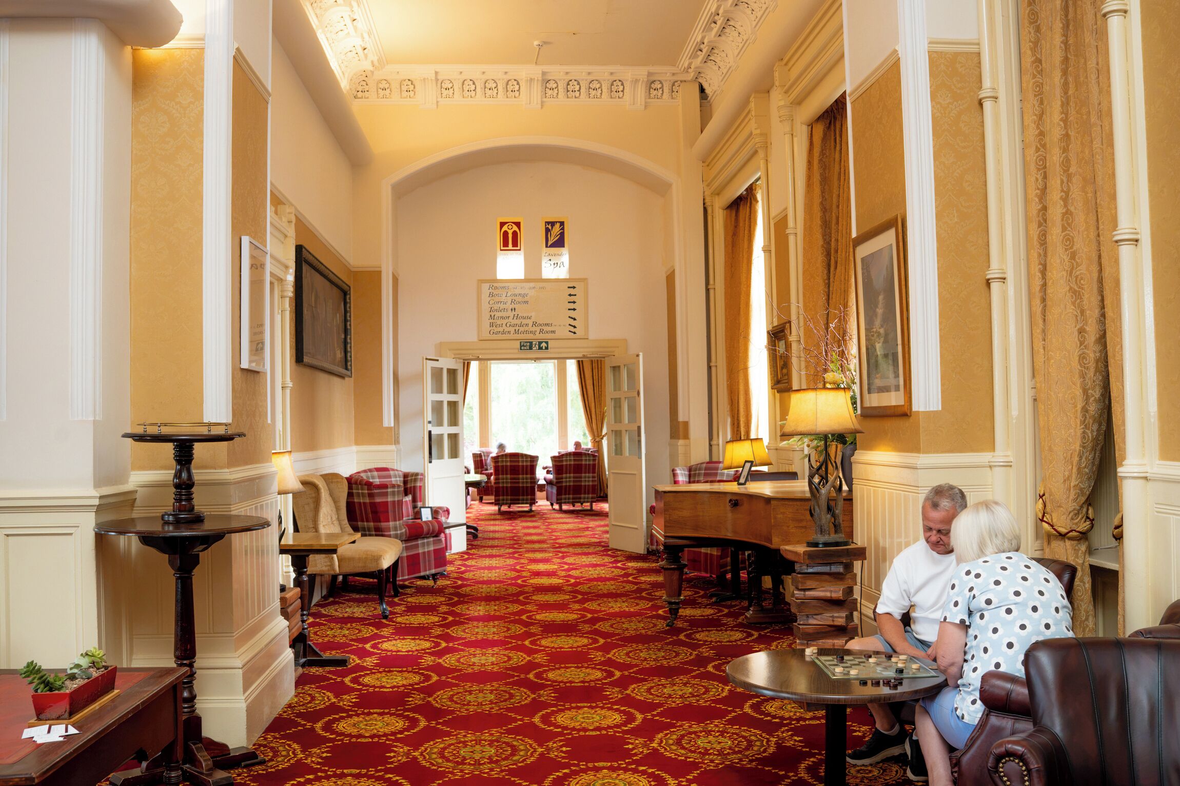 Un hall d’hôtel avec un tapis rouge, des espaces aménagés avec des sièges, un comptoir d’accueil et des panneaux d’orientation.