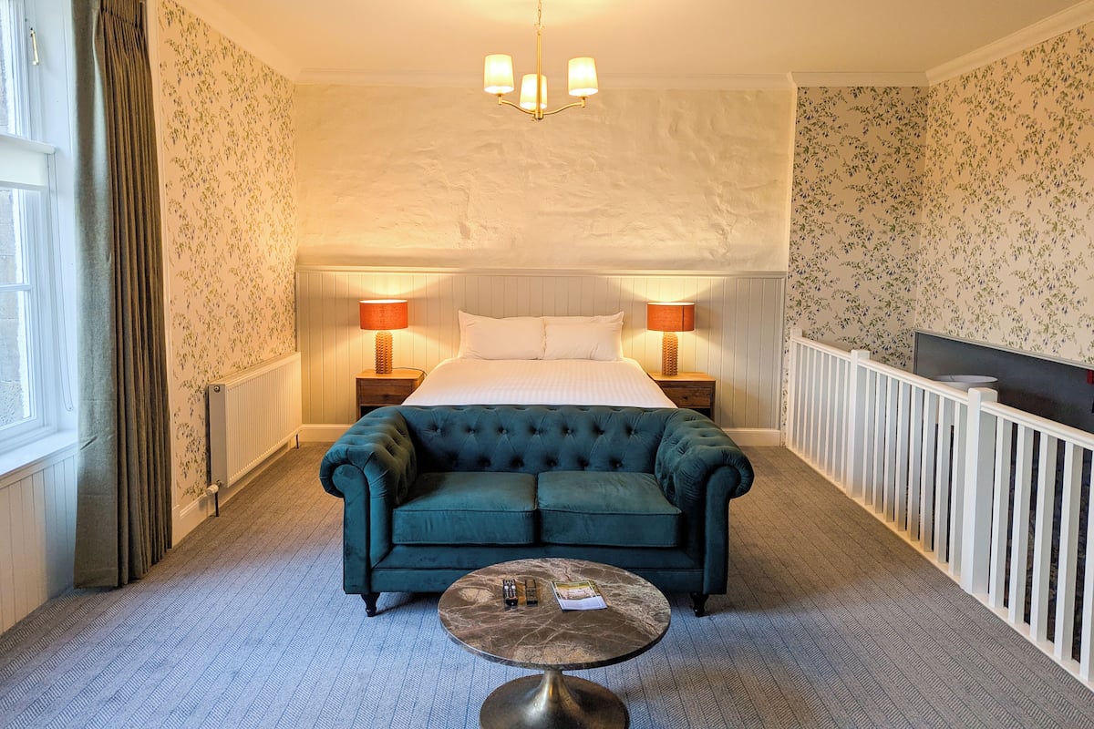 Une chambre d’hôtel avec un grand lit, deux tables de chevet avec des lampes, un canapé vert, une table basse ronde et une vue sur l’extérieur.