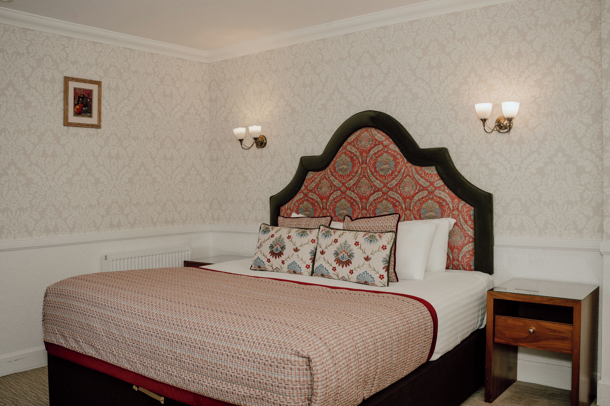 Ein Schlafzimmer mit einem gemusterten Kopfteil, einem Bett mit roter und beiger Bettdecke, zwei Wandleuchten und einem Nachttisch mit Schublade.