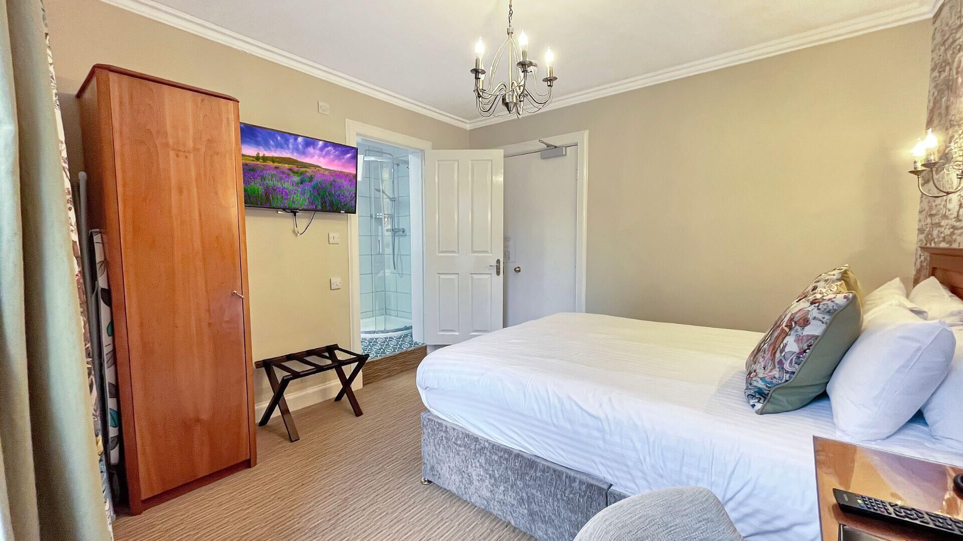 A bedroom with a bed, a wooden wardrobe, a small table, a shower area, and a painting on the wall.
