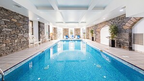 An indoor swimming pool with stone walls and tiled flooring, surrounded by lounge chairs and potted plants.