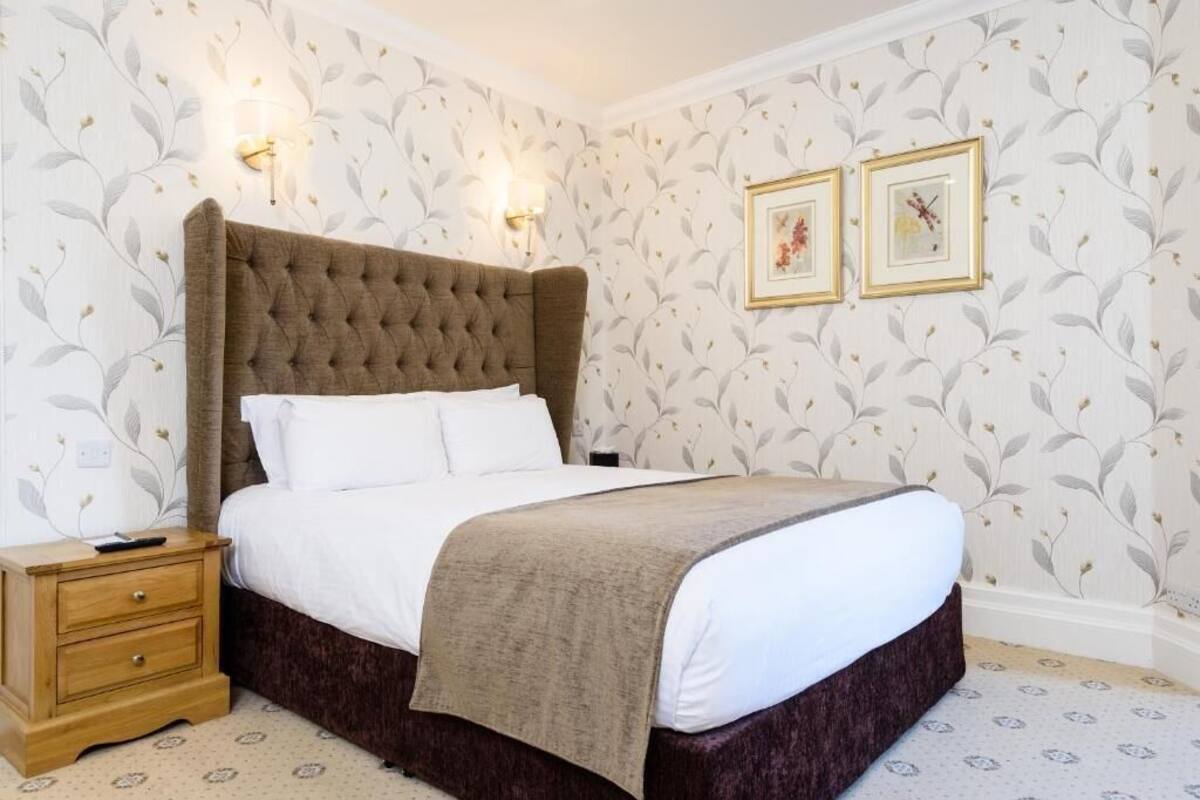 Une chambre à coucher avec une tête de lit capitonnée, une table de chevet en bois dotée de tiroirs, un lit avec du linge de lit blanc et un plaid beige, ainsi que deux appliques murales.