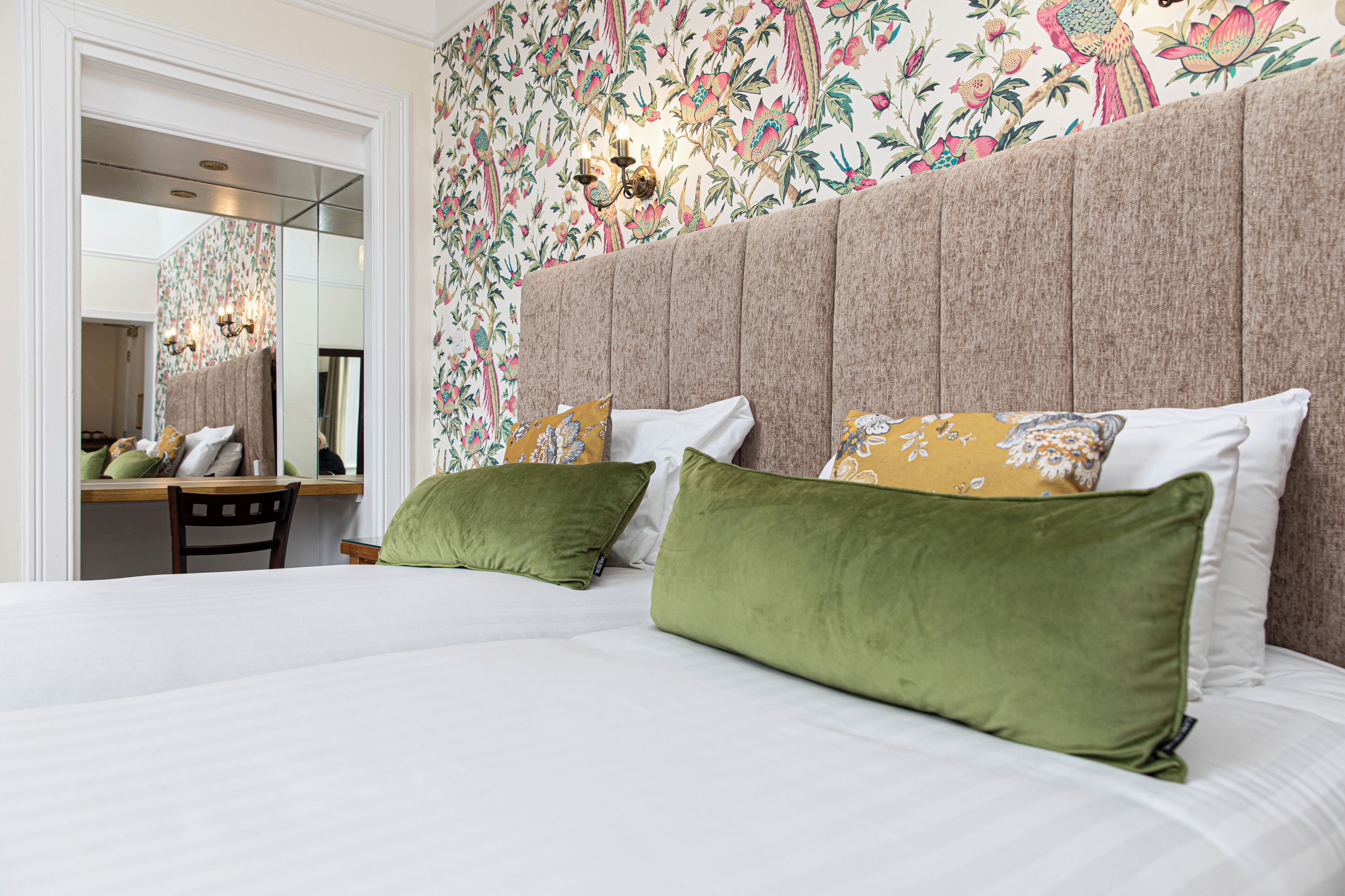 A bedroom with a large bed, green and yellow pillows, a mirror, and a floral wallpaper.