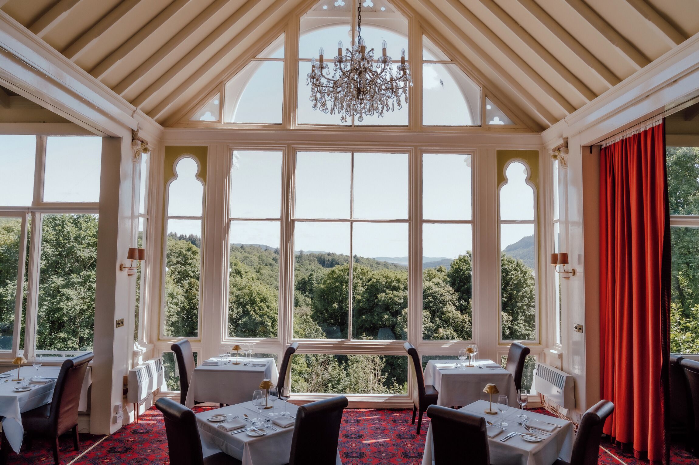 A dining area with tables set for four, large windows offering a view of a forested landscape, and a chandelier hanging from the ceiling.