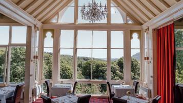 Un comedor con mesas dispuestas para cuatro, ventanas amplias que ofrecen vistas a un paisaje boscoso y una araña colgando del techo.