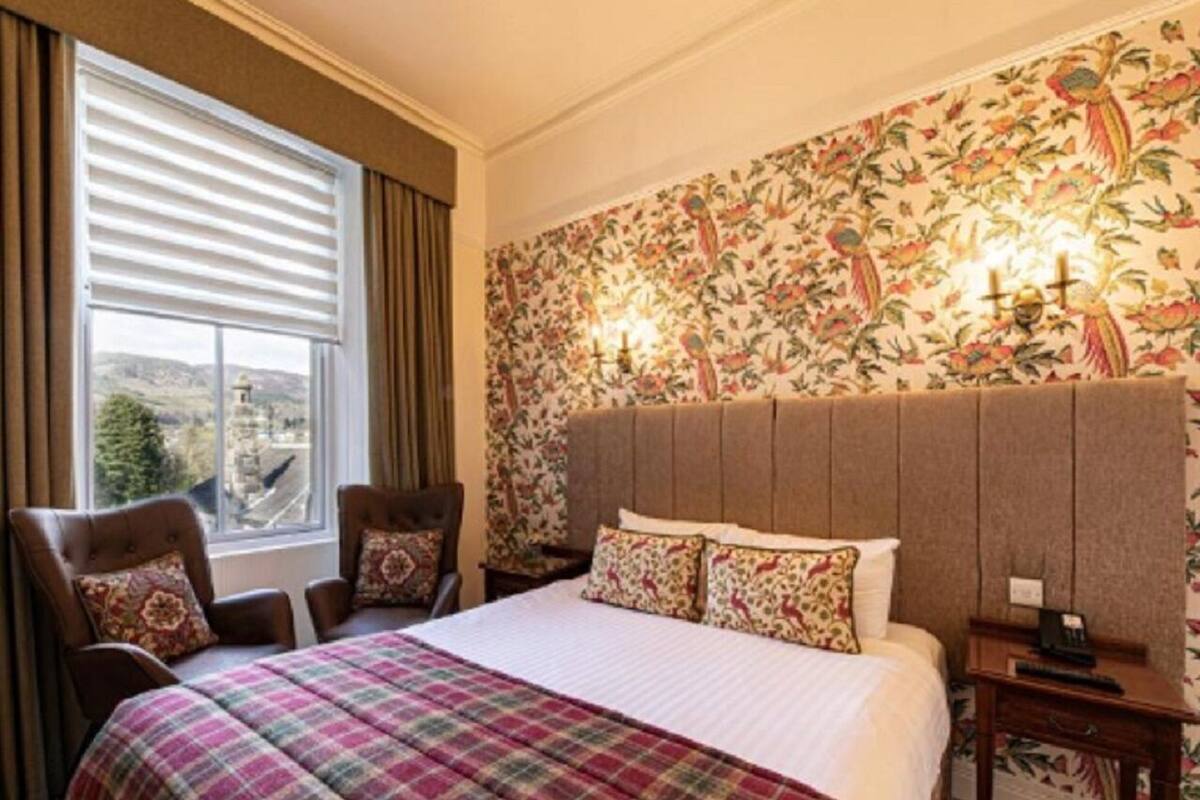 Une chambre d’hôtel avec un grand lit, deux fauteuils, une table de chevet et une fenêtre avec des rideaux.