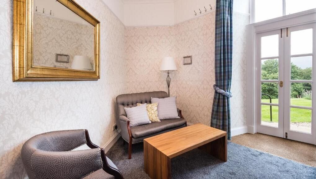 A room with a sofa, a chair, a wooden table, a mirror, a lamp, and a view through a door.