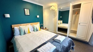 Ein ordentlich bezogenes Bett mit vielen Kissen, ein Nachttisch mit Lampe, ein großer Spiegel und ein offener Kleiderschrank.