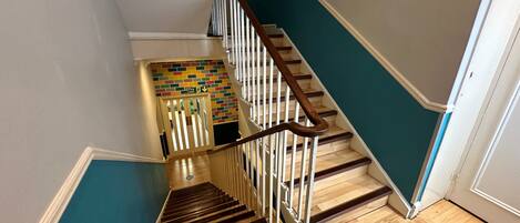 A staircase with wooden steps and white railings, leading to a landing with a colorful wall and a fire exit sign.