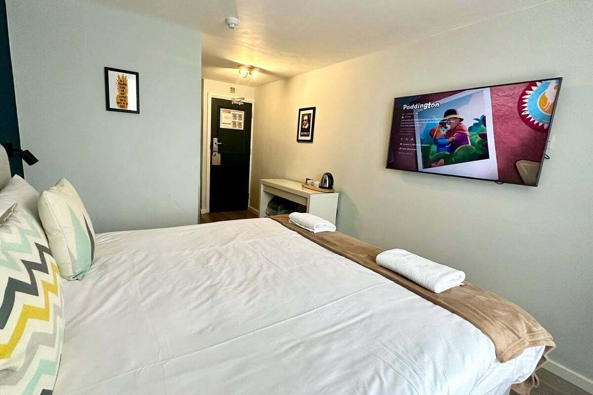 A hotel room with a bed, pillows, a TV, and a small table with a kettle.