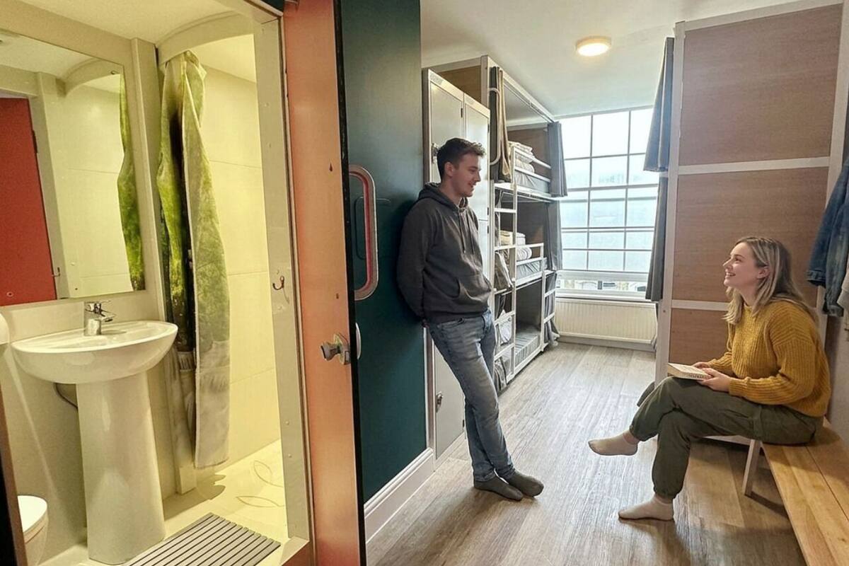 A bathroom with a sink, shower, and a door leading to a room with a bench and a person sitting.