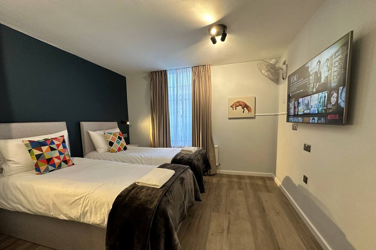 A hotel room with two beds, a large flat-screen TV mounted on the wall, a ceiling fan, and a window with curtains.