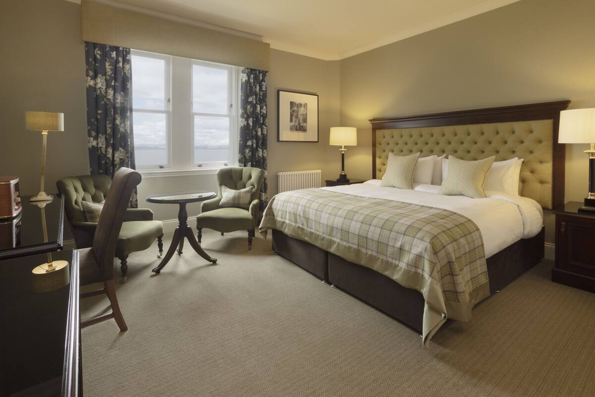 A hotel room with a large bed, two armchairs, a small table, and a window with curtains.