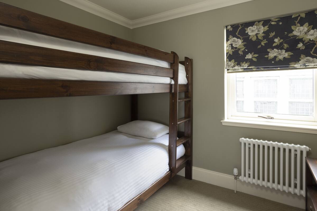 A bunk bed with white bedding, a radiator below a window with floral curtains, and a plain wall.