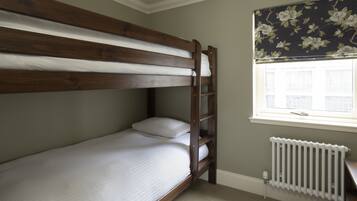 Una cama litera con ropa de cama blanca, un radiador debajo de una ventana con cortinas florales y una pared lisa.